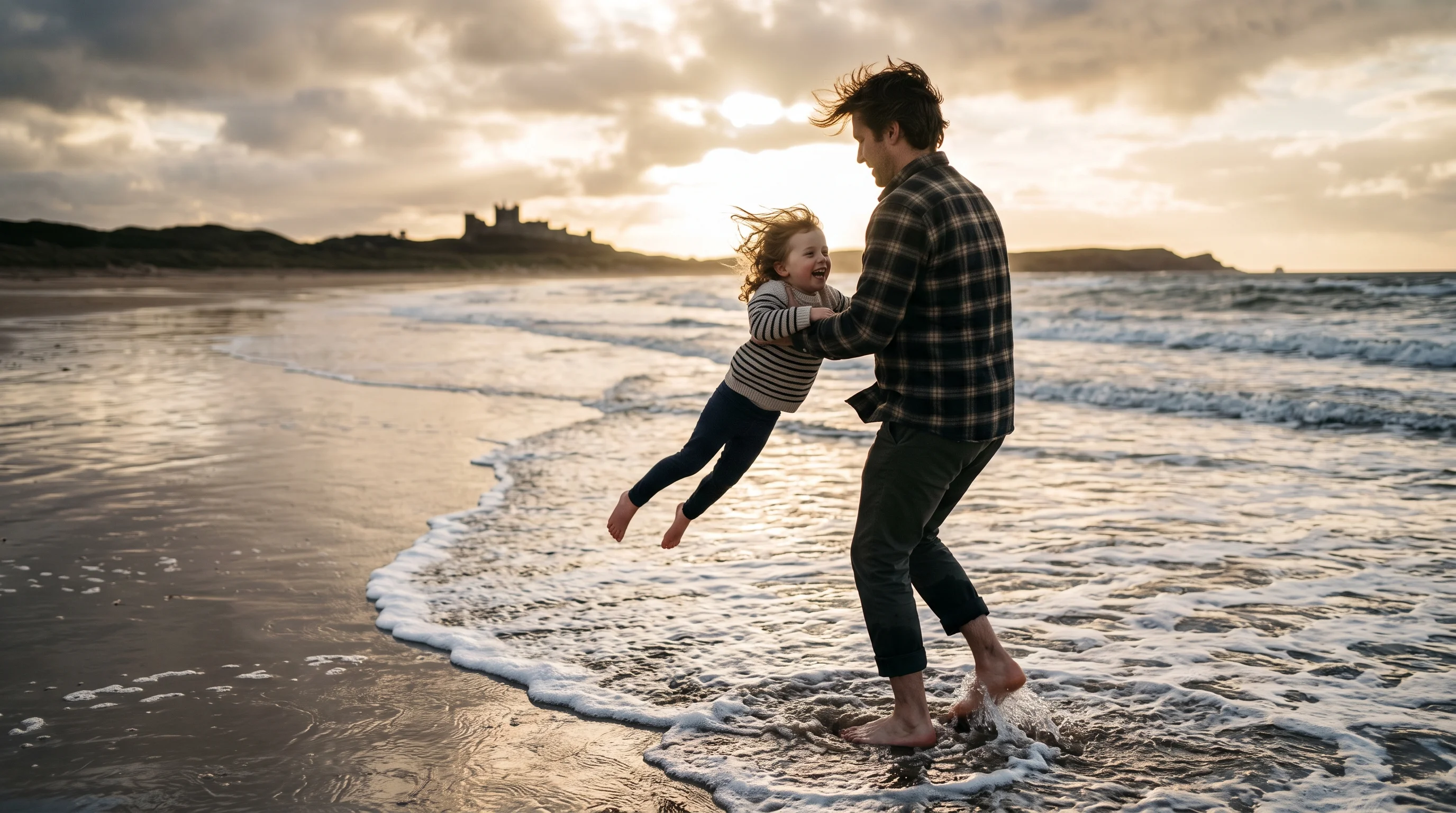A parent swings a child high into the air at the water's edge, their shadows stretching across the wet sand