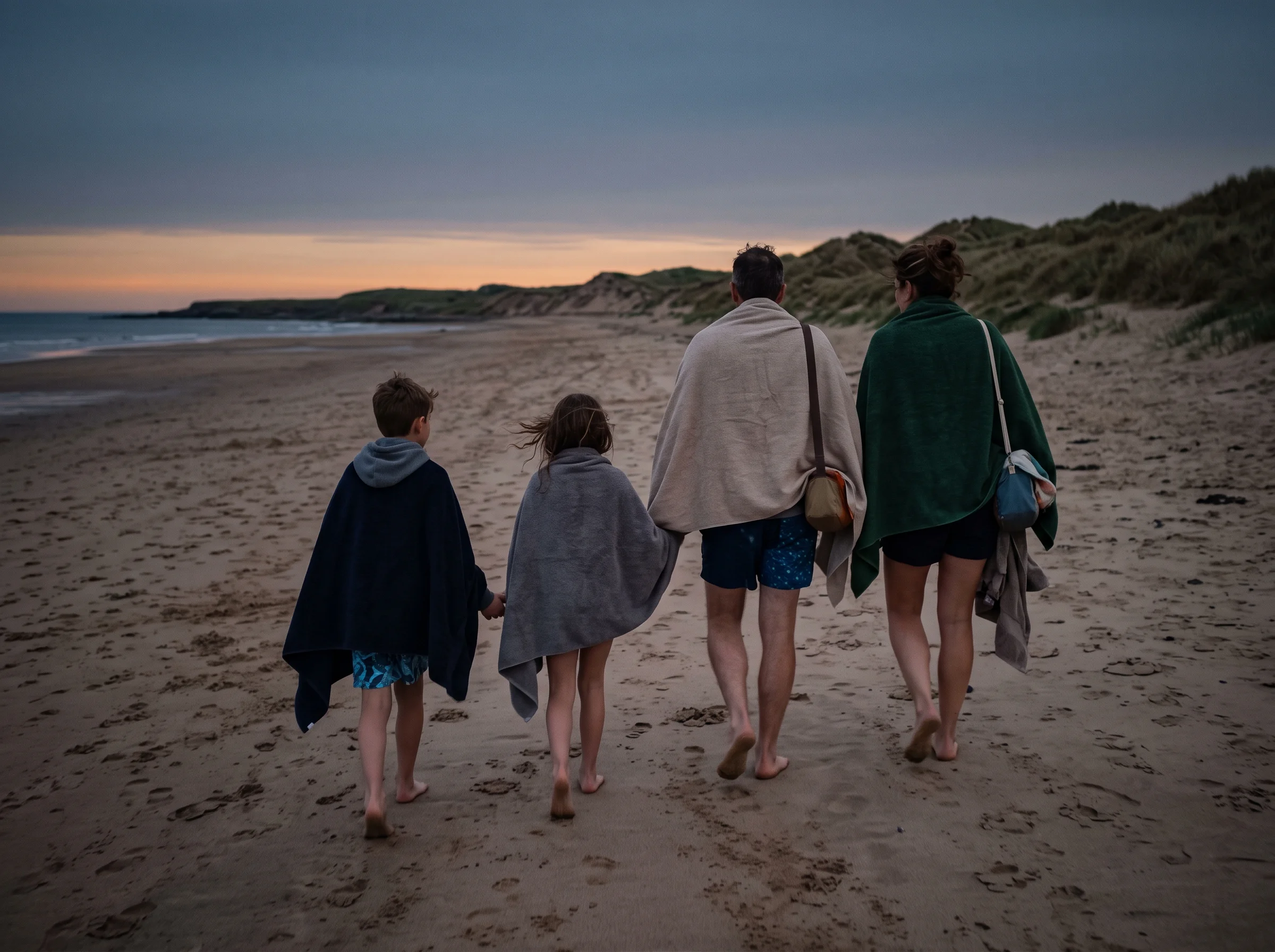 A family of four walks away from the camera along a sandy beach at dusk, all wrapped in towels.