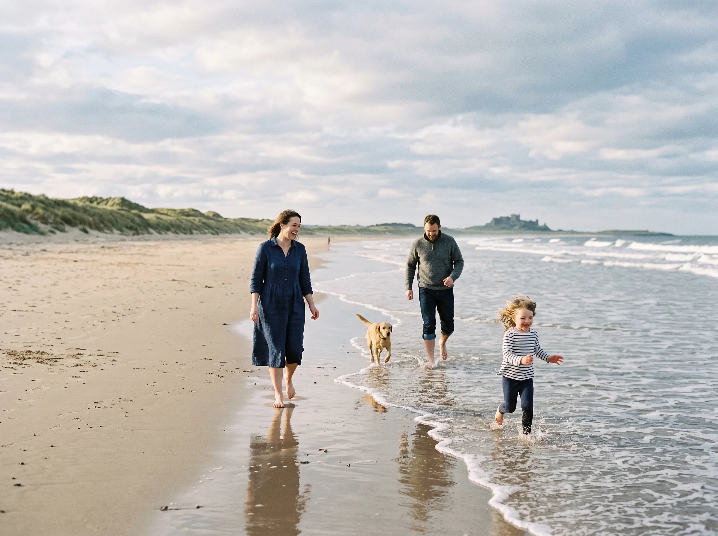 A family with a child, a man, a woman, and a dog walking along a sandy beach near the tide line.