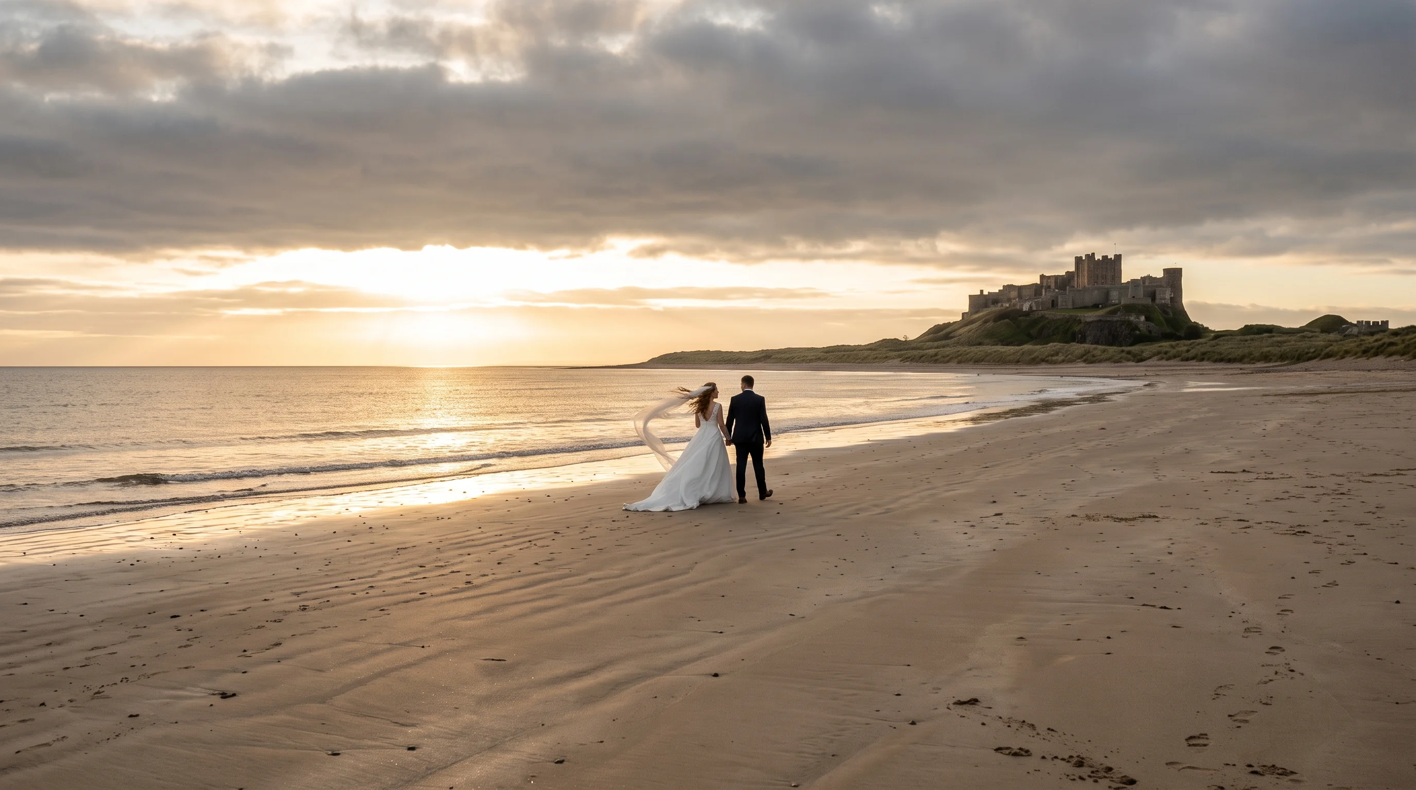A couple stand small on a wide windswept beach at golden hour, her veil blown sideways, with a castle on the headland behind them