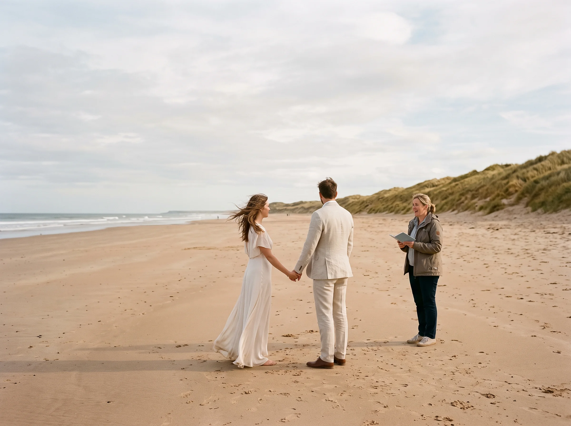 A couple stand alone on a wide beach with a celebrant, dunes rising behind them