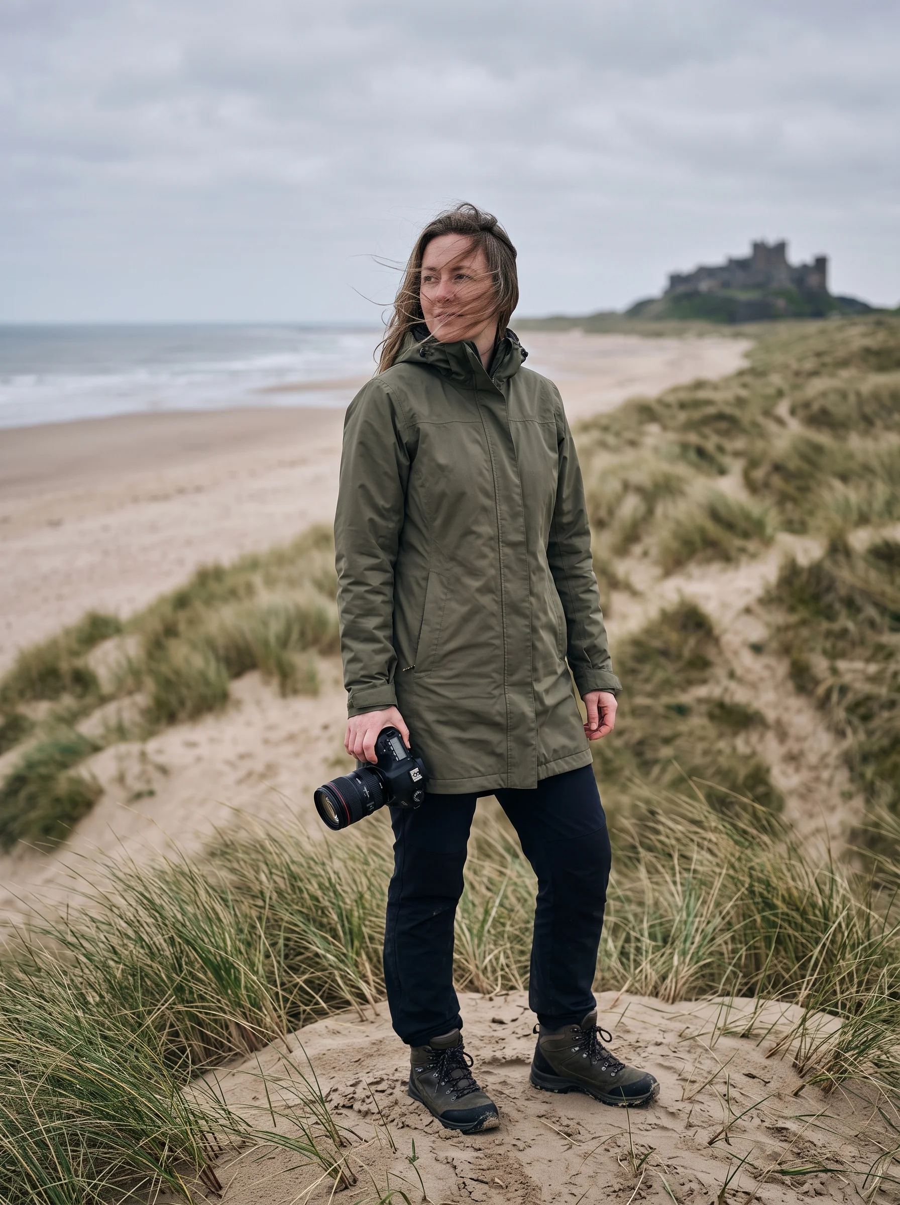 A photographer in a windproof coat stands on a dune with her camera lowered, hair blown across her face, half-smiling at something off to the side