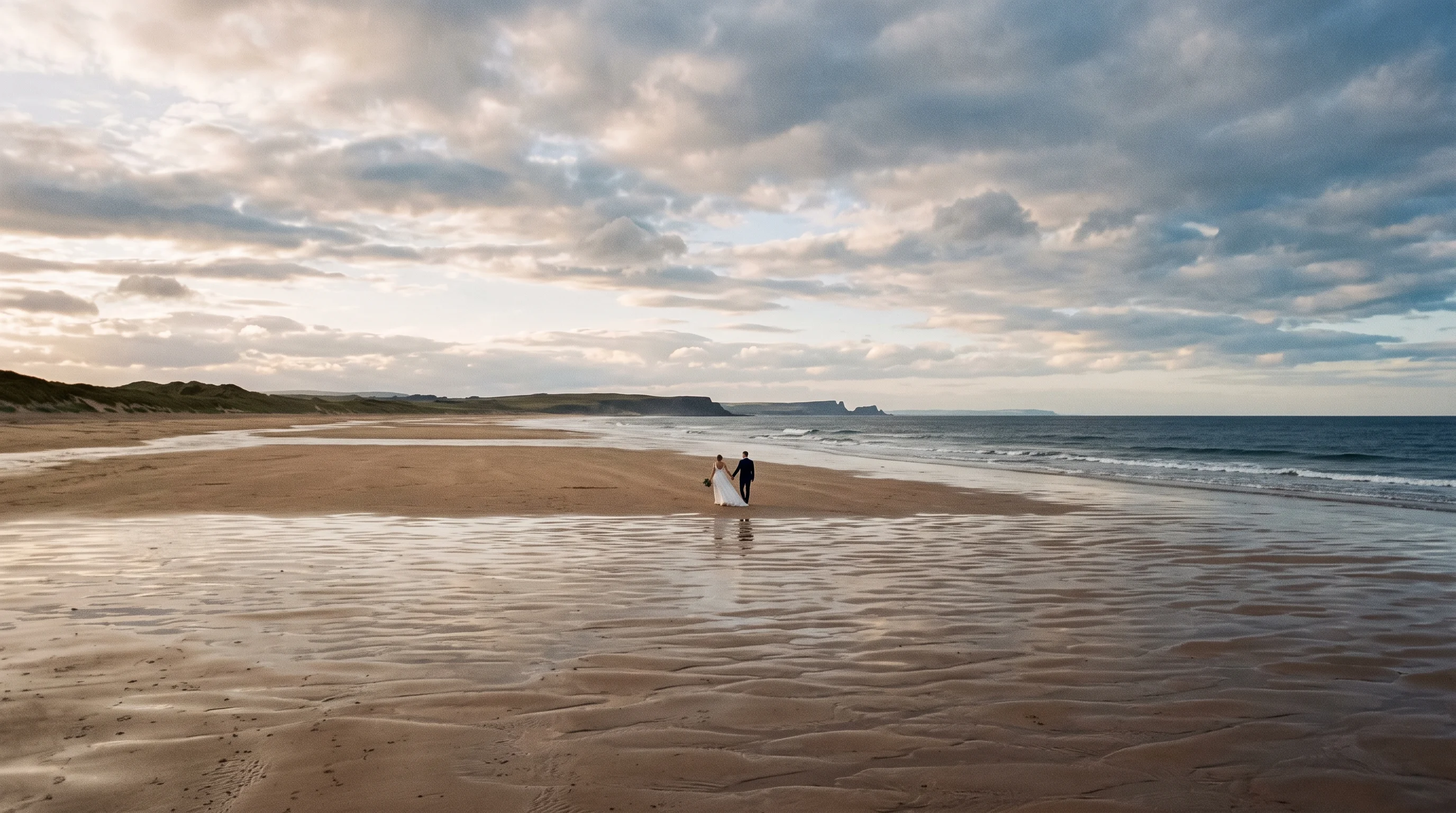 A couple walk as two small figures along a wide empty beach at low tide under a huge windy sky