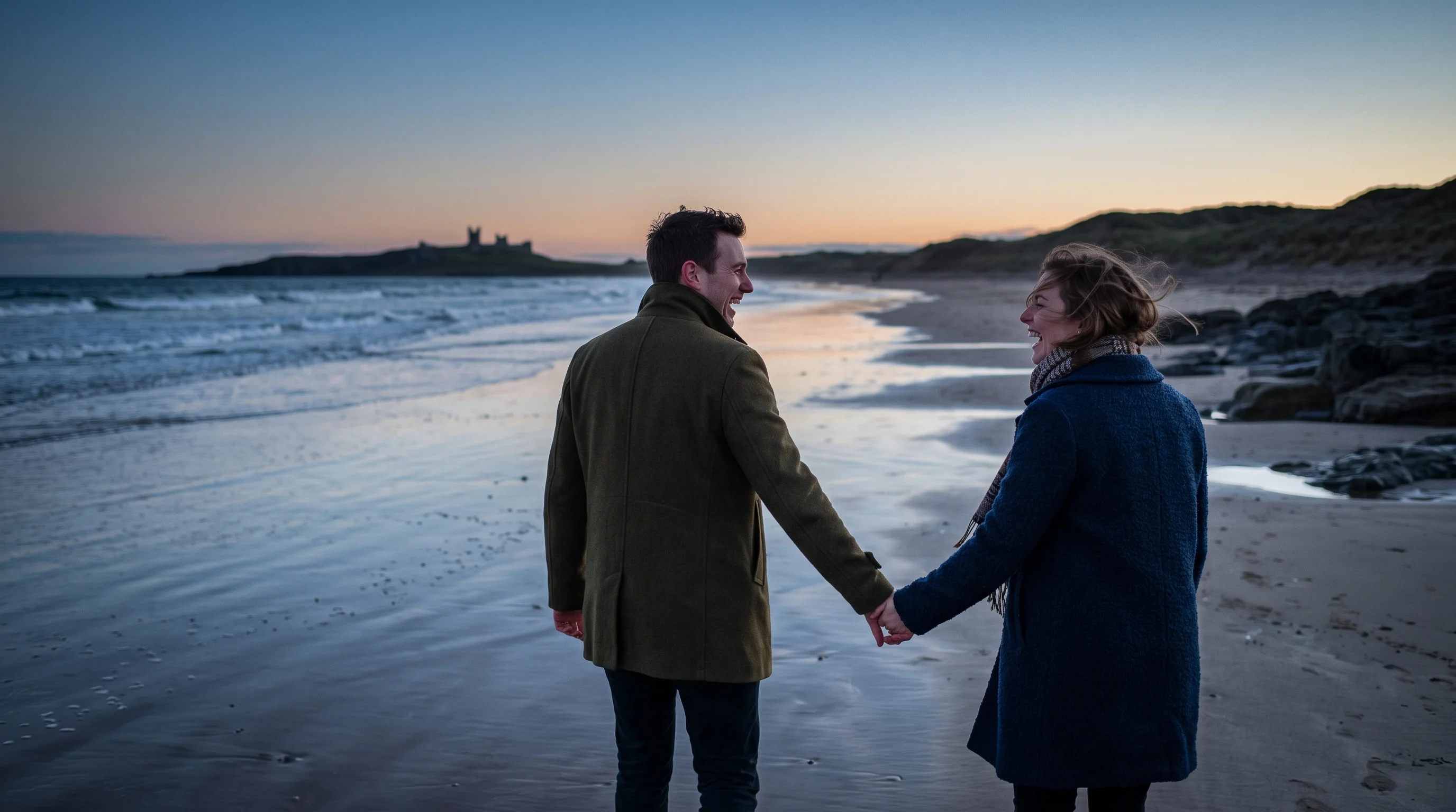 A couple in coats walk away along the shoreline at dusk, laughing into the wind