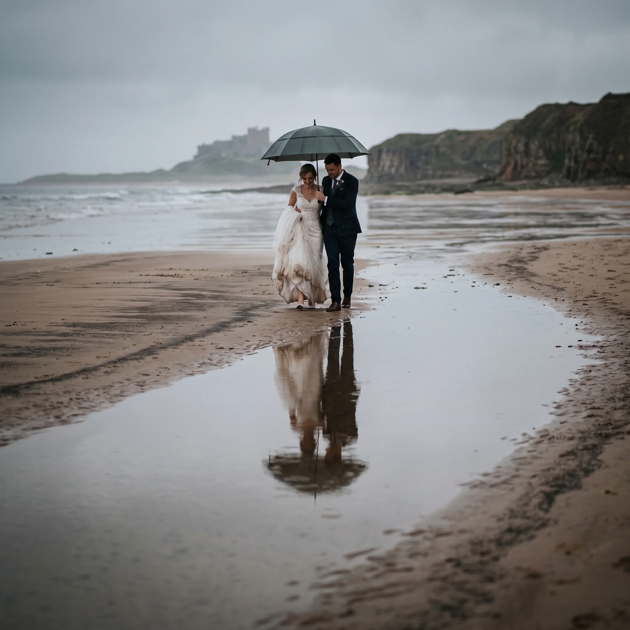 A couple under one umbrella stand on mirror-wet sand, their reflection beneath them in the rain