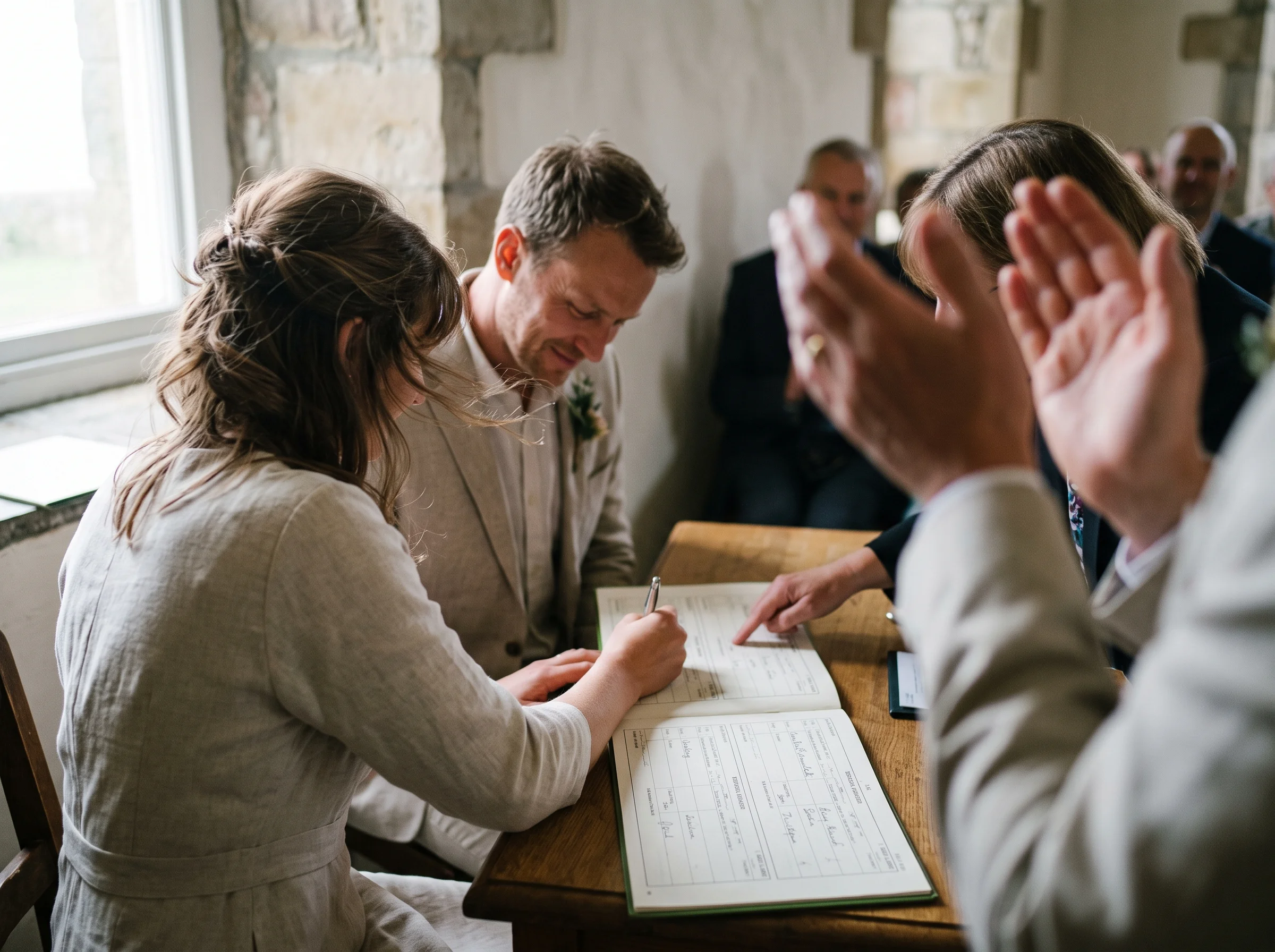 A couple sign the register at a small table while two witnesses' hands clap blurred at the edge of the frame