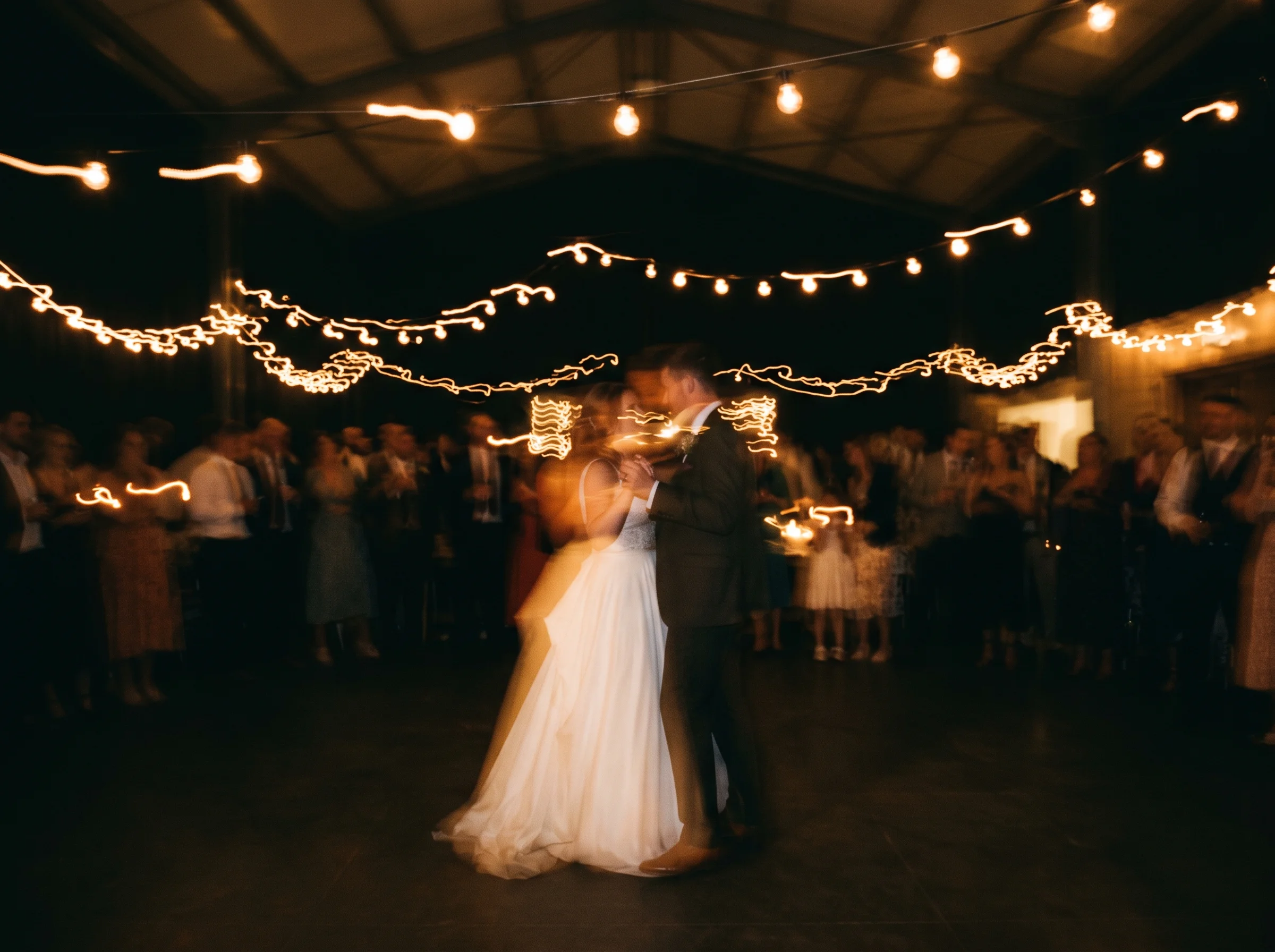 A couple turn in their first dance, blurred under festoon lights, guests soft in the dark around them