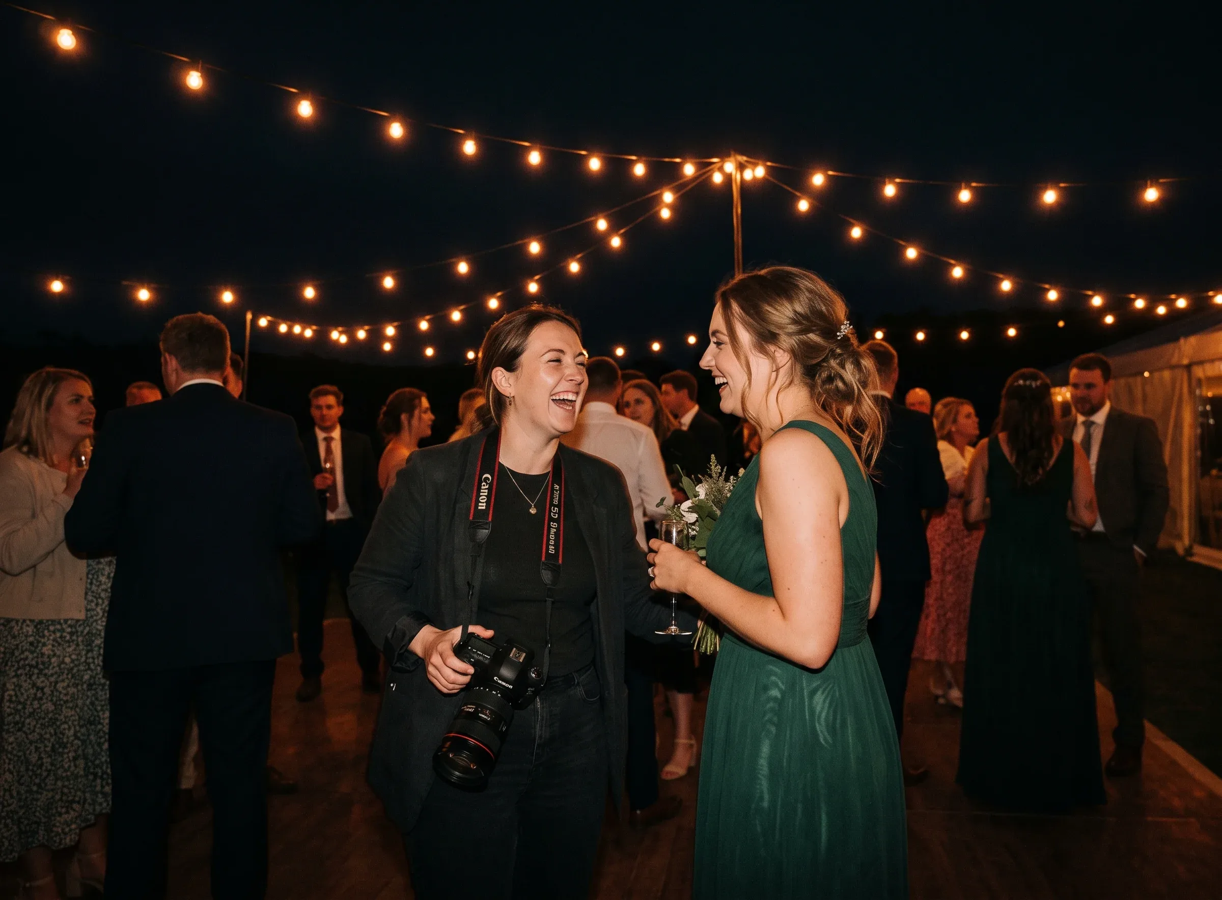 A guest's snapshot of a photographer laughing with a bridesmaid under festoon lights in the evening