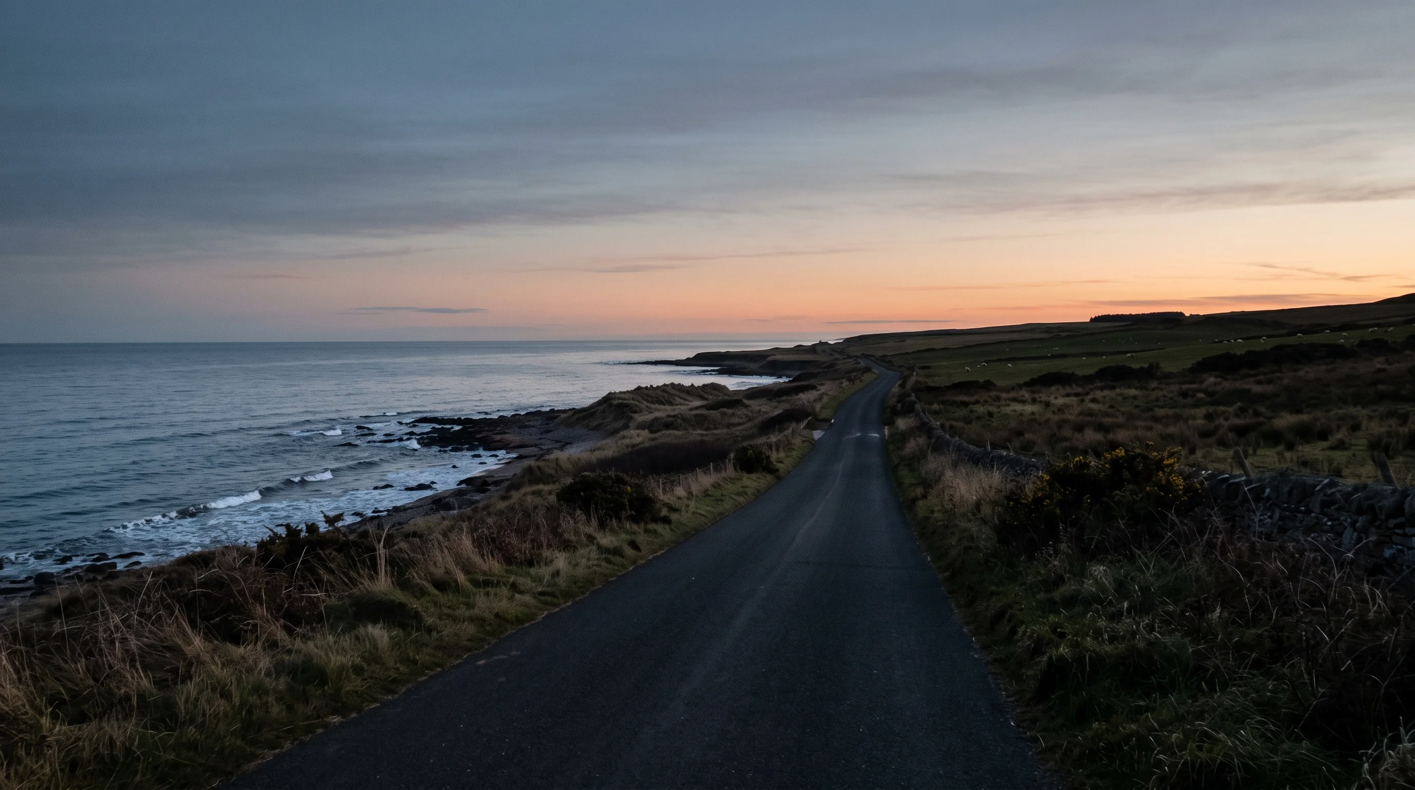 An empty coast road at dusk with the sea on one side, fields on the other and a band of amber light on the horizon