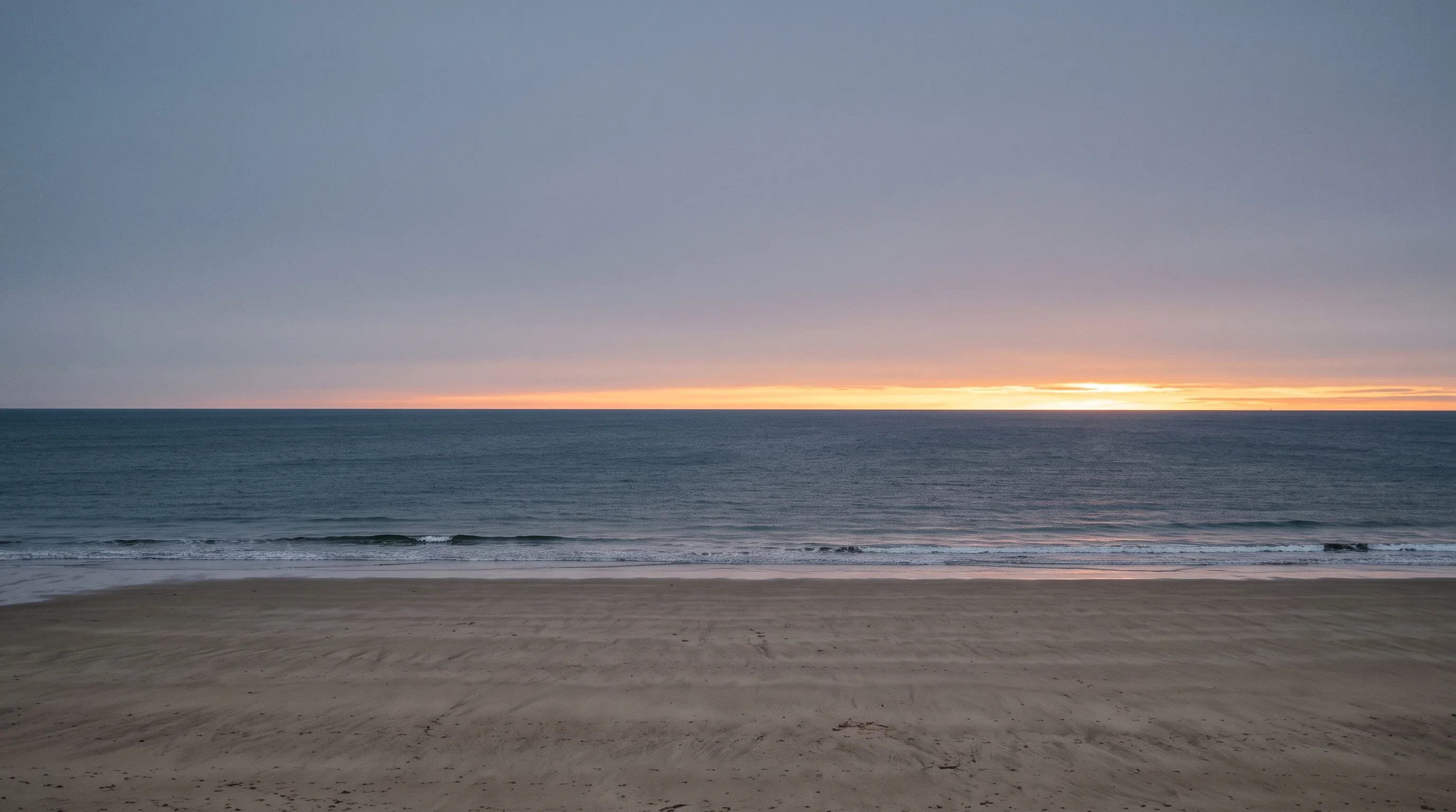 An empty beach at dawn, wet sand reflecting a thin band of light along a low horizon